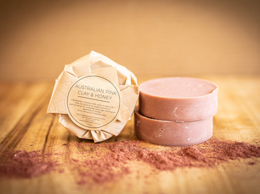 A round bar of soap packed in brown paper is propped next to two unwrapped soaps. The bars are pink and round. They sit on a timber board and there is pink clay powder sprinkled in front. The label on the soap pack says Australian pink clay and honey, by Honey and Glow
