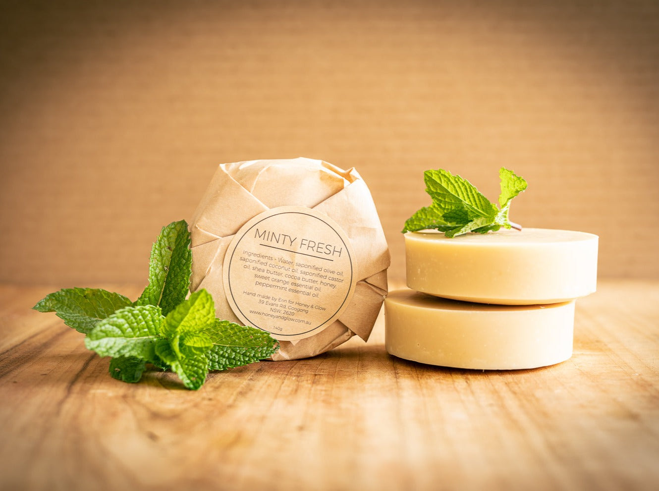 A pack of round soap wrapped in brown paper is propped next to two unwrapped round bars of soap. The label on the pack says Minty Fresh by Honey and Glow. There are sprigs of mint to the left and on top of the bars.