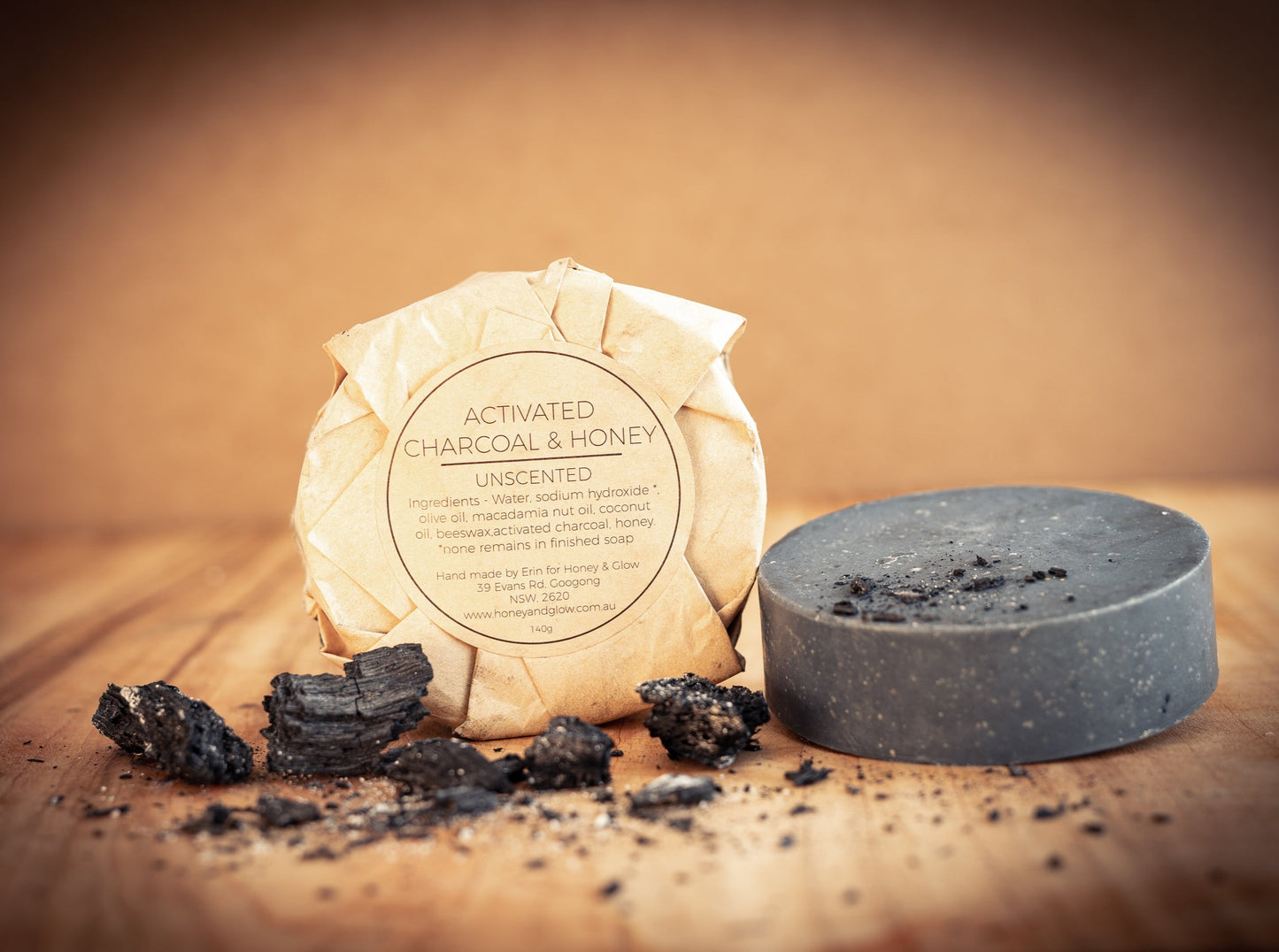 A wrapped pack of round soap is propped next to an unwrapped soap bar. The unwrapped bar is round and black. There are pieces of charcoal around. The label on the pack says Activated Charcoal and Honey, unscented, by Honey and Glow.